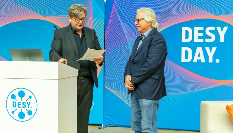 ein Moderator überreicht eine Urkunde an einen Mann mit weißen Haaren, Brille und Schnauzbart